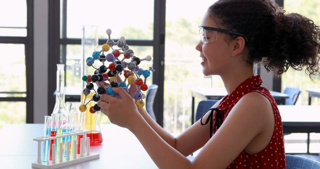 Young girl in school laboratory studying a molecular model while wearing safety goggles. Ideal for educational websites, science program promotions, and STEM learning content. Demonstrates hands-on learning and academic curiosity in a classroom setting.