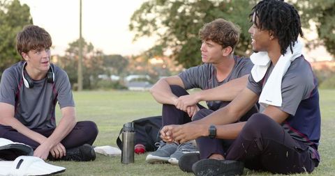 Diverse male teammates resting on grass after practice talking, hydrating, bonding