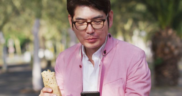 Man enjoying a wrap while using a smartphone outdoors, wearing a pink blazer and glasses. Ideal for representing casual dining, mobile technology usage, and outdoor relaxation. Suitable for promoting healthy eating habits, urban lifestyle, and technological engagement.