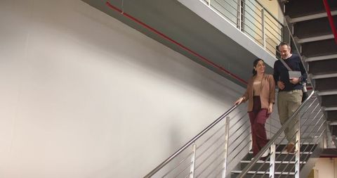 Diverse coworkers descending industrial staircase holding rail carrying bag and folder