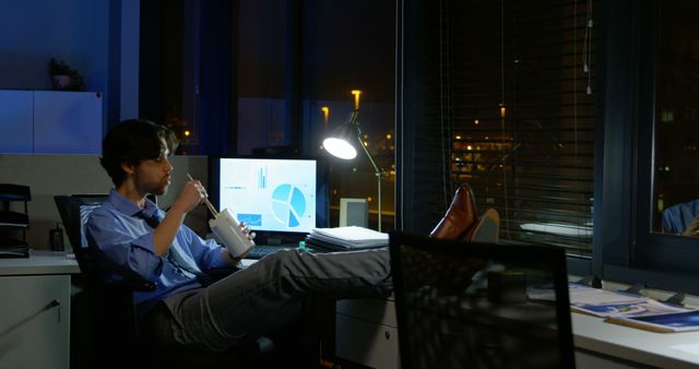 Businessman taking a break working late at night in an office. Sitting relaxing with cup and feet on desk. Computer screens displaying charts and graphs visible in background. Reflected city lights seen through window. Useful for illustrating high-pressure work environments, corporate lifestyles, work-life balance themes, or productivity challenges.