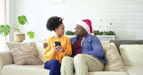 Couple relaxing on couch celebrating cozy holiday with smartphone and Santa hat