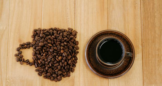 Creative arrangement of coffee beans in the shape of a cup next to a filled brown coffee cup on a wooden background. Perfect for illustrating coffee culture, morning routines, or caffeine energy themes. Great for use in blogs, social media, or advertisements for coffee shops.