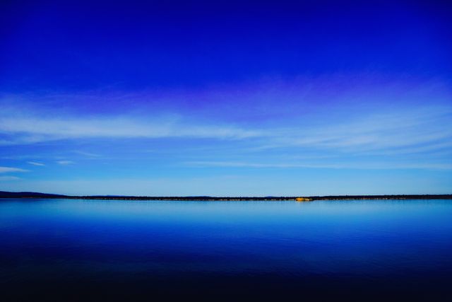 This image captures a stunning blue sky reflecting over a calm lake during sunset. The serene setting with a clear horizon and gentle water creates a tranquil and peaceful atmosphere, making it ideal for use in travel advertisements, relaxation themes, or nature blog posts. The bright yet soft colors of the sunset offer a sense of calmness and beauty.