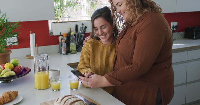 Two friends are sharing a joyful moment in a bright kitchen while checking a smartphone. Fresh breakfast items, including juice and bread, are on the counter. This stock photo can be used for concepts such as bonding, social media, lifestyle, and everyday life.