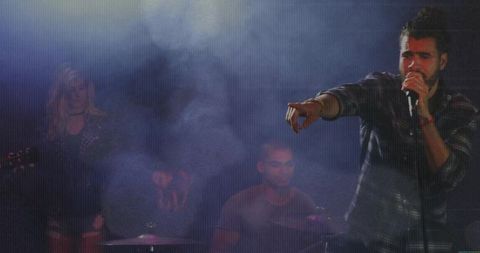 Lead vocalist pointing and singing on foggy backlit stage with drummer and guitarist