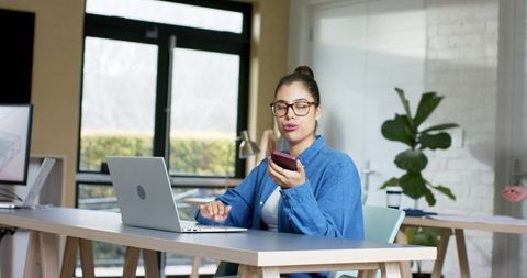 Young woman working from coworking desk using laptop and voice commands on smartphone