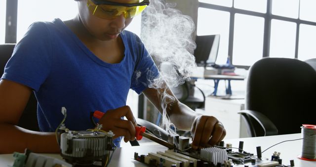 A young African American woman wearing safety goggles is soldering components on an electronic board, with smoke rising from her work, indicating active soldering. Her focused expression and the equipment suggest she is skilled in electronics or engineering.
