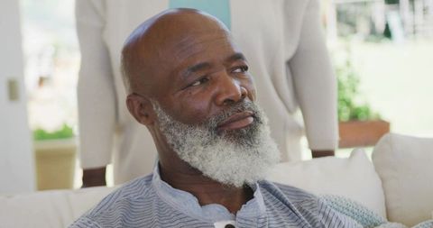 Senior man sitting on sofa gazing while partner standing behind in bright living room