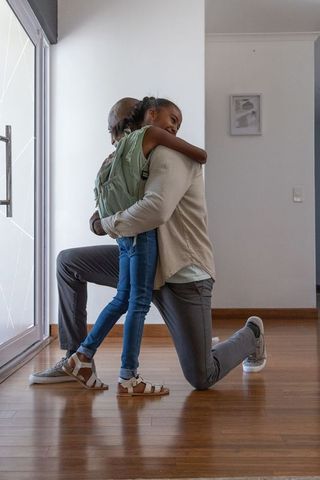 Kneeling father hugging teen daughter at front door with backpack — homecoming bond