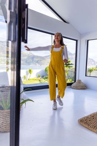 Senior woman strolling through sunlit coastal living space in yellow overalls