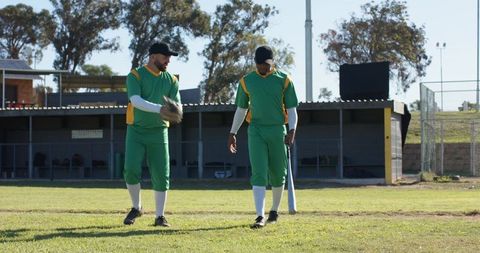 Two teammates walking on baseball field holding bat and glove near dugout at golden hour