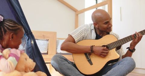 Father playing acoustic guitar with african american daughter holding teddy in sunlit bedroom