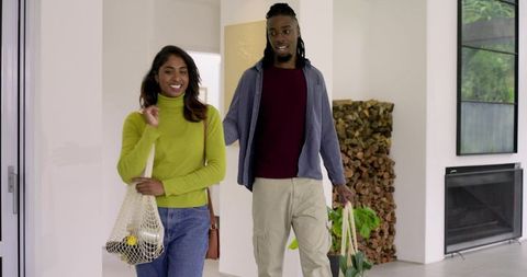Diverse couple walking into modern home carrying groceries, tote bag and houseplant