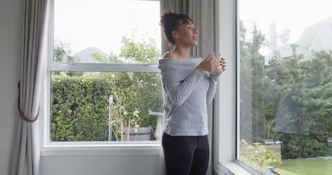 Woman standing by window holding mug, enjoying calm morning in cozy off-shoulder sweater