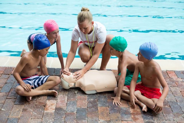 Female lifeguard demonstrating CPR techniques to a group of children by the poolside. Ideal for use in educational materials, safety training programs, summer camp promotions, and swimming pool safety campaigns.