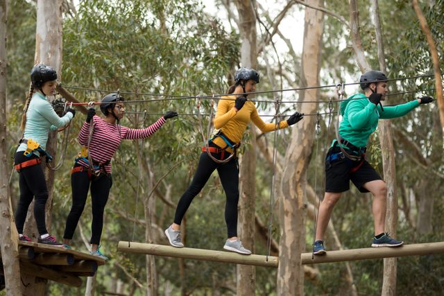 Group of friends participating in a zip line adventure in a forest. They are wearing safety gear including helmets and harnesses, and are balancing on wooden planks. This image can be used for promoting outdoor activities, team-building events, adventure tourism, and recreational parks.