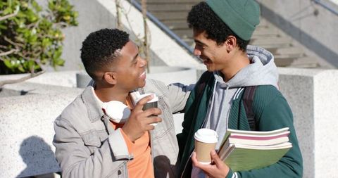 College friends sharing coffee, laughing and chatting on campus stairs while carrying books