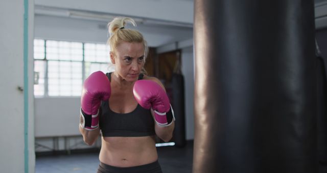 Middle-aged woman workouts in gym, training with punching bag, exemplifies strength and focus in fitness routine. Use for advertising fitness programs, women's health, self-defense classes, or boxing training.