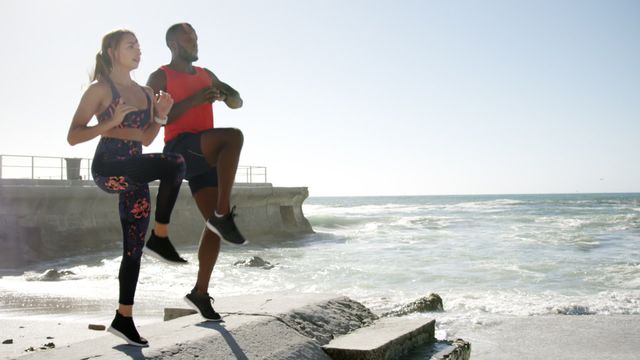 This video captures a multi ethnic couple performing high knees on a beach. The two are dressed in athletic wear, focusing on their fitness routine under a clear sky next to the ocean waves. Perfect for promoting an active and healthy lifestyle, outdoor workout programs, fitness classes, personal training, sportswear advertising, and summer fitness campaigns.