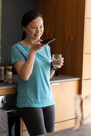 Asian woman speaking into smartphone while holding glass of water in modern kitchen