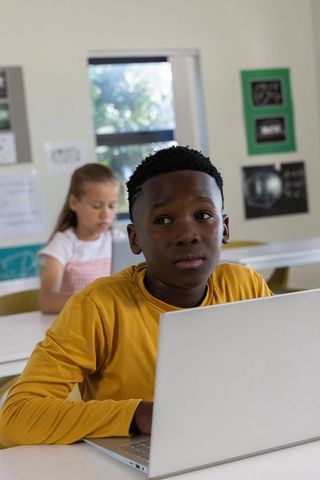 Focused black student using laptop while classmate writing in sunlit classroom learning