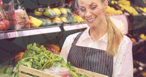 Smiling produce clerk lifting crate of fresh greens while checking stock via network overlay