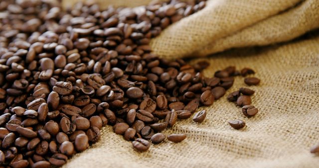 Roasted coffee beans are scattered across a burlap surface, with a sack partially filled with more beans in the background, with copy space. The image evokes the rich aroma and the initial stages of coffee preparation.