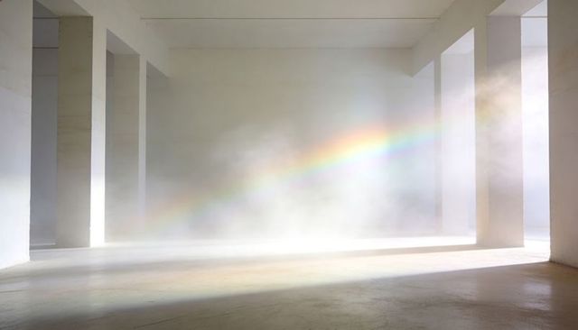 Diagonal rainbow band cutting through mist-filled concrete hall, sunlight flooding pillars