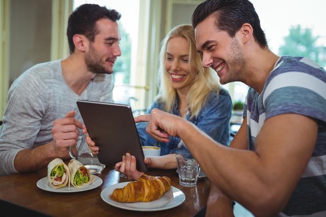 Group of friends sitting at a table in a cafe, enjoying breakfast and using a digital tablet. They are smiling and engaging with each other, creating a warm and friendly atmosphere. Ideal for use in advertisements for cafes, technology products, or social apps. Can also be used in lifestyle blogs or articles about socializing, friendship, and modern dining experiences.