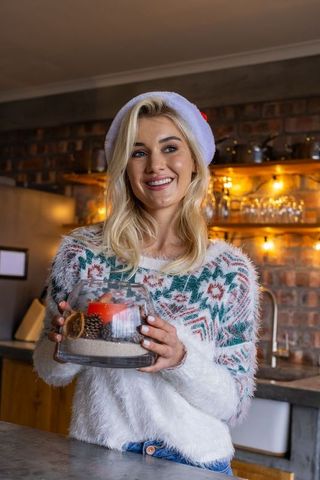 Smiling woman in santa hat holding festive glass centerpiece in cozy kitchen