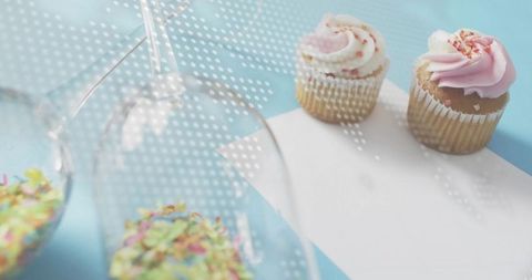Serving pastel cupcakes with pink frosting and sprinkles on blue tabletop with inverted glasses