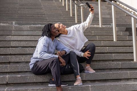 Diverse women in athleisure sitting on concrete steps taking joyful selfie outdoors