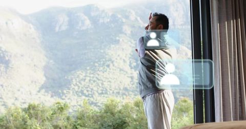 Asian man stretching by floor-to-ceiling window overlooking mountains with holographic ui