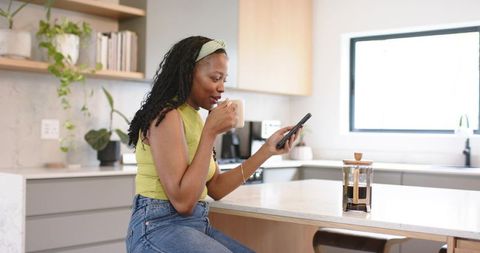 African american woman sipping coffee and checking smartphone on modern kitchen island
