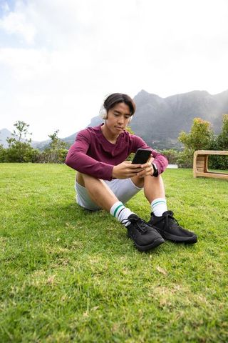 Asian man sitting on grassy lawn listening to music and checking smartphone