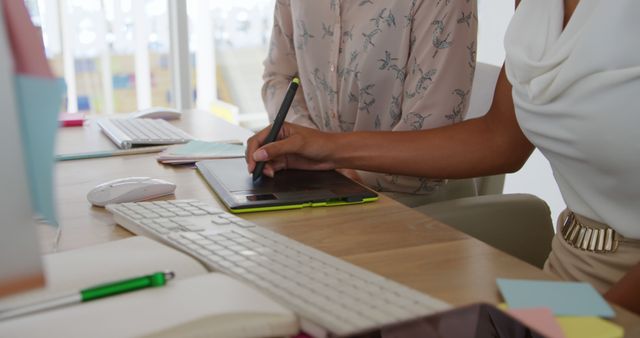 Two women are collaborating on work at a desk, one using a digital tablet. They are in a modern office environment, suggesting professionalism and creativity. This can be used for topics related to business, teamwork, collaboration, technology use in offices, professional settings, and creative workspaces.