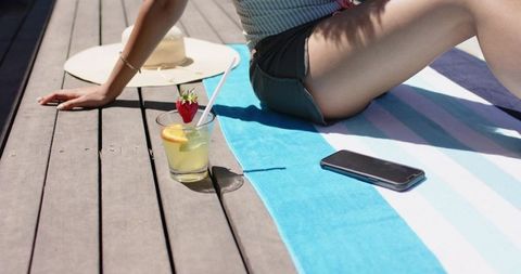 Woman relaxing on striped beach towel tapping smartphone beside lemonade and sun hat