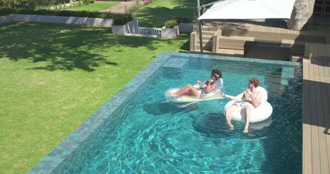 Couple relaxing on inflatable floats in turquoise backyard pool on sunny summer afternoon
