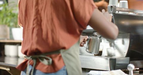 Female barista steaming milk at espresso machine holding stainless pitcher for latte art