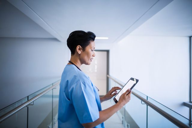 Female nurse in blue uniform using digital tablet in a hospital corridor. Ideal for illustrating modern healthcare, medical technology, and professional healthcare workers. Suitable for use in healthcare websites, medical blogs, and promotional materials for hospitals and clinics.