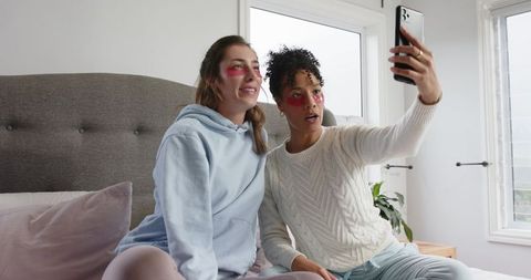 Two friends taking selfie on bed wearing under-eye patches pastel bedroom self-care moment