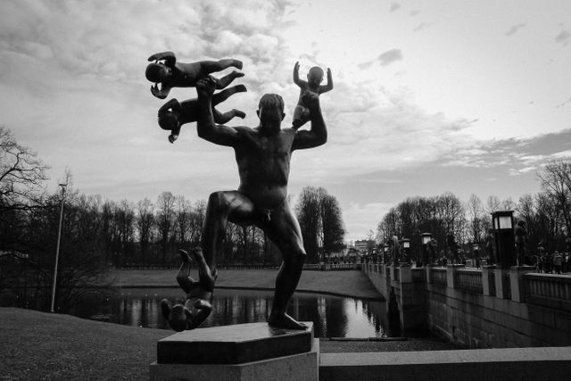This black and white image showcases a dynamic metal sculpture in a park, depicting a figure energetically lifting multiple smaller figures. The detailed craftsmanship and fluid movement make it an engaging piece of public art. The background includes a serene park landscape with a calm body of water, a bridge featuring lamps, and barren trees. It captures the art's intricacy and the park's tranquil atmosphere. Ideal for use in historical studies, art appreciation materials, public art promotion, or highlighting cultural landmarks.