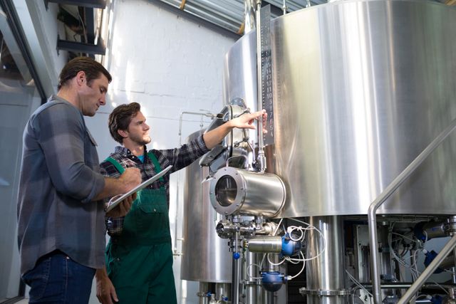 Two workers in a brewery discussing the brewing equipment. One worker is pointing at a gauge while the other takes notes on a clipboard. The setting is industrial with large stainless steel tanks and machinery. This image can be used for themes related to manufacturing, teamwork, industrial processes, and professional collaboration.