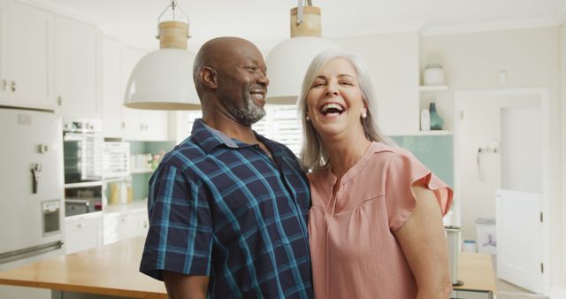 Senior couple laughing together in modern kitchen. Ideal for use in advertisements, blog posts about family life, retirement, happiness, home life, and relationship advice. Highlights joyful moment, emphasizing strong bond and love in a relaxing home environment.