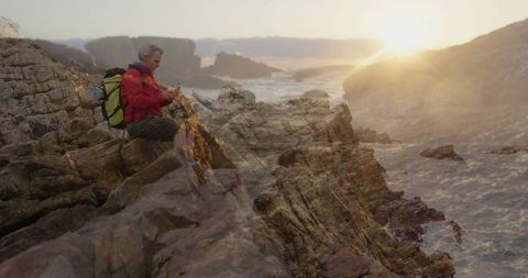 Hiker with Backpack Checking Smartphone on Coastal Rocks at Sunset