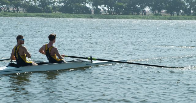 Two male rowers are training on a double scull boat on a lake. Wearing athletic gear, they focus on their rowing technique and coordination. This image can be used to promote teamwork, sports events, fitness activities, and rowing competitions.