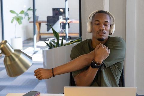 African american man stretching at desk wearing headphones while working on laptop in coworking spac