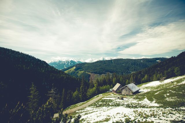 Panoramic view of an Alpine landscape featuring traditional mountain huts nestled in a secluded valley. Snow covers the ground partially, with dense forests and majestic mountains in the background under a vast sky. Ideal for themes related to nature, wilderness, tranquility, outdoor activities, and travel destinations.