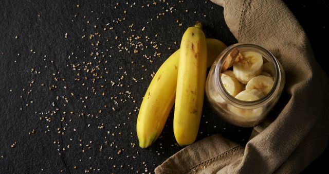 Fresh bananas alongside a jar of sliced bananas rest on a dark surface scattered with crumbs, with copy space. Bananas are a nutritious fruit, high in potassium and fiber, often included in a healthy diet.